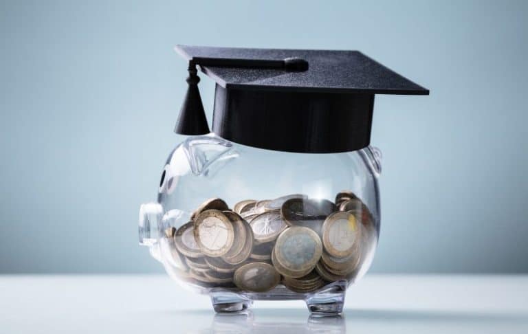 A clear piggy bank filled with coins is topped with a black graduation cap, symbolizing saving money for education or student financial planning.