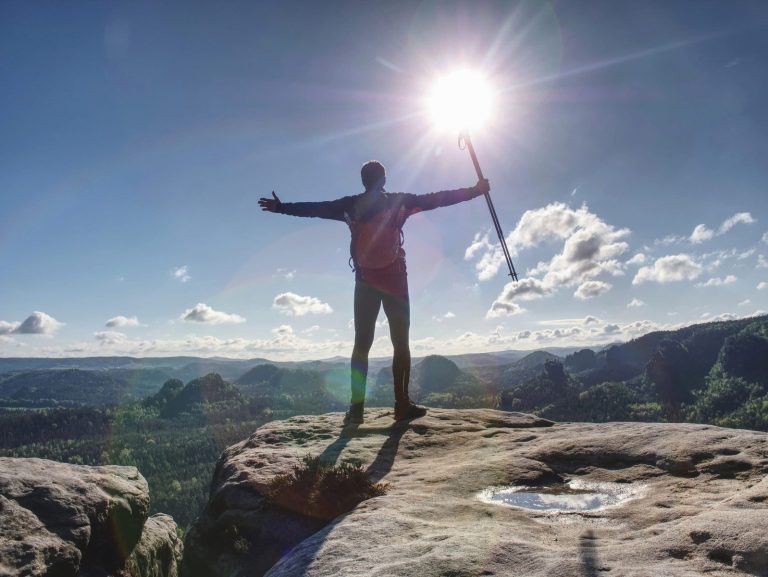 A person stands on a rocky cliff with arms outstretched, holding trekking poles, facing the sun—like the triumphant moment described in a Coalition App essay. Below stretch mountains, trees, and a bright blue sky dotted with clouds.
