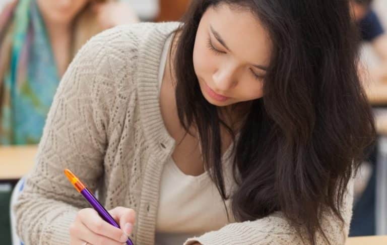 A young woman with long dark hair, wearing a beige sweater, is sitting at a desk and writing with a pen in a classroom setting. Other students are blurred in the background.