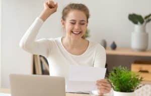 A young woman sits at a desk, smiling joyfully with her fist raised in celebration while holding a piece of paper. A laptop and a green potted plant are in front of her.