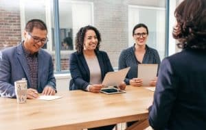Three people in business attire sit at a table, smiling and holding papers, facing another person whose back is to the camera, suggesting a job interview or meeting in a modern office setting.
