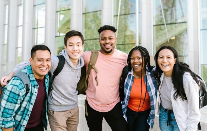Five young adults, smiling and standing close together with arms around each other, pose outdoors in front of a modern glass building. They appear happy and casual, wearing backpacks and casual clothing.