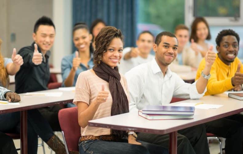 A diverse group of students sitting at desks in a classroom, smiling and giving thumbs up towards the camera. Notebooks and textbooks are visible on the desks.