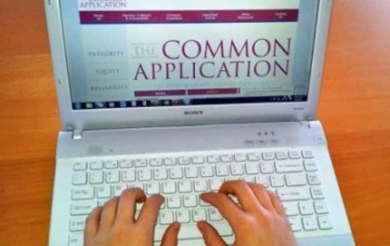 A person’s hands typing on a white laptop keyboard with a screen displaying the Common Application website for college admissions. The background is a wooden surface.