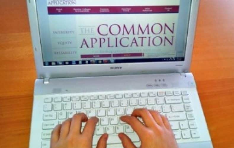 A person’s hands typing on a white laptop keyboard with a screen displaying the Common Application website for college admissions. The background is a wooden surface.