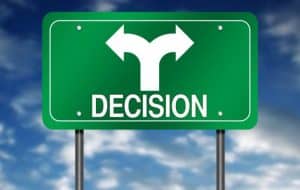 Green road sign with the word “DECISION” and a white split arrow pointing left and right, symbolizing a choice or two possible directions, against a blue sky with clouds.