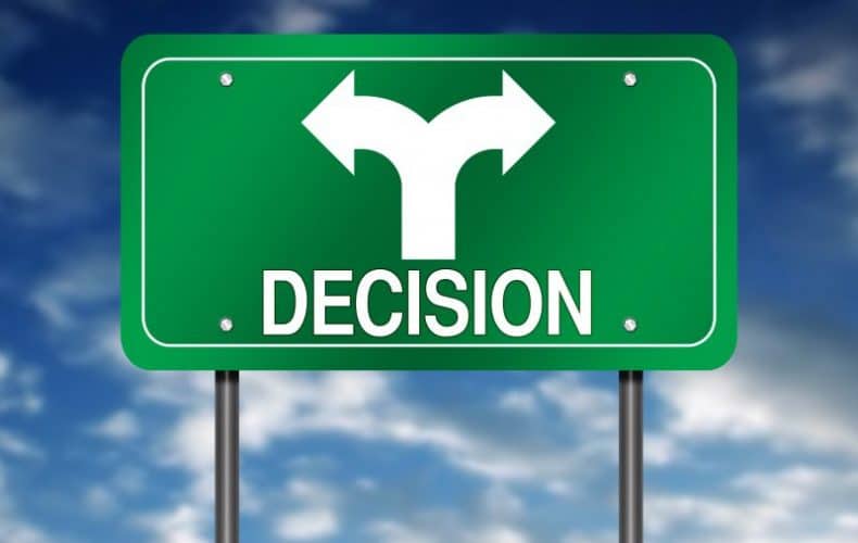 Green road sign with the word “DECISION” and a white split arrow pointing left and right, symbolizing a choice or two possible directions, against a blue sky with clouds.