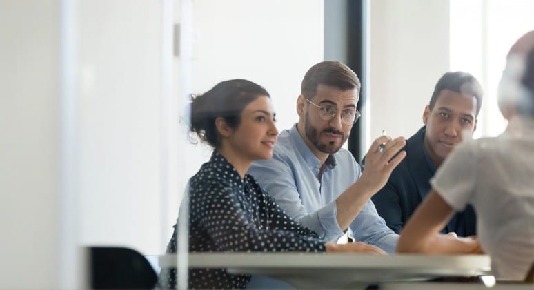 Three people sit at a table in a meeting room, engaged in conversation with a fourth person who is partially visible. One man gestures while speaking, and the group appears attentive and focused.