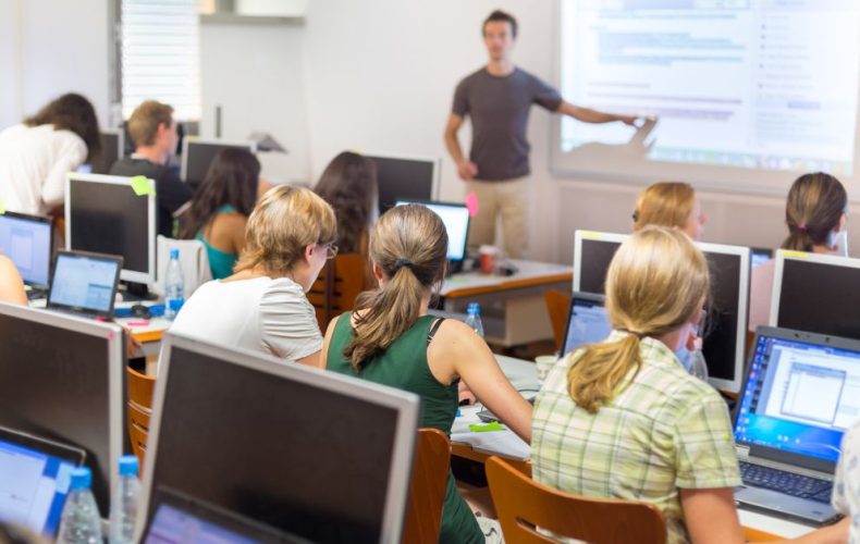 students sitting in classroom and teacher is standing while poiting towards screen