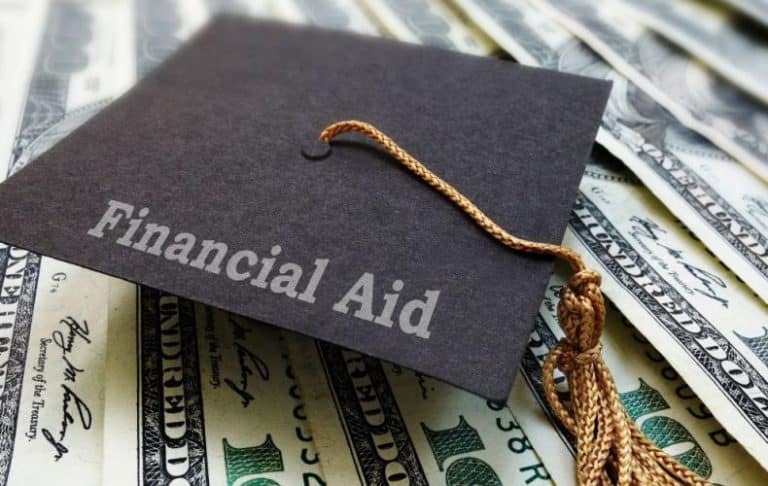 A graduation cap labeled Financial Aid rests on top of several spread out U.S. one hundred dollar bills.