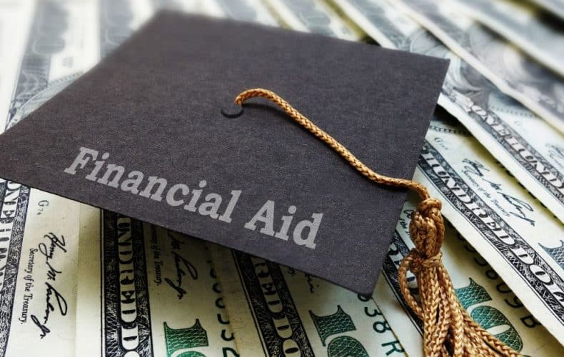 A graduation cap labeled Financial Aid rests on top of several spread out U.S. one hundred dollar bills.