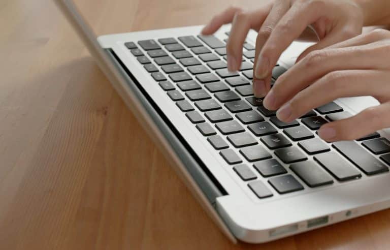 Close-up of a persons hands typing on a laptop keyboard on a wooden surface. The laptop is silver, and only a portion of the device and hands are visible.