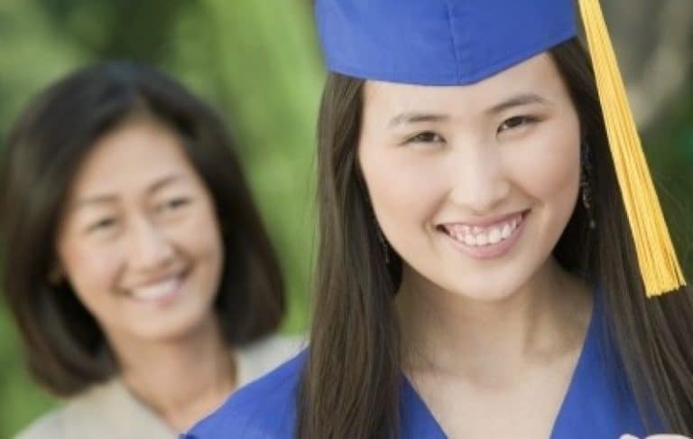 A young woman in a blue graduation cap and gown smiles at the camera, with a smiling older woman standing in the background outside.
