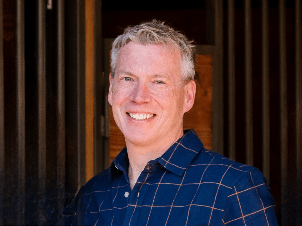 A man with short gray hair smiles at the camera, wearing a blue shirt with an orange grid pattern, standing in front of a wooden background.
