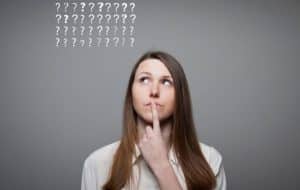 A woman with long brown hair looks up thoughtfully with her finger on her lips. Above her head are rows of question marks on a gray background, suggesting she is thinking or unsure.