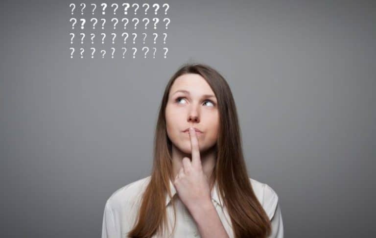 A woman with long brown hair looks up thoughtfully with her finger on her lips. Above her head are rows of question marks on a gray background, suggesting she is thinking or unsure.