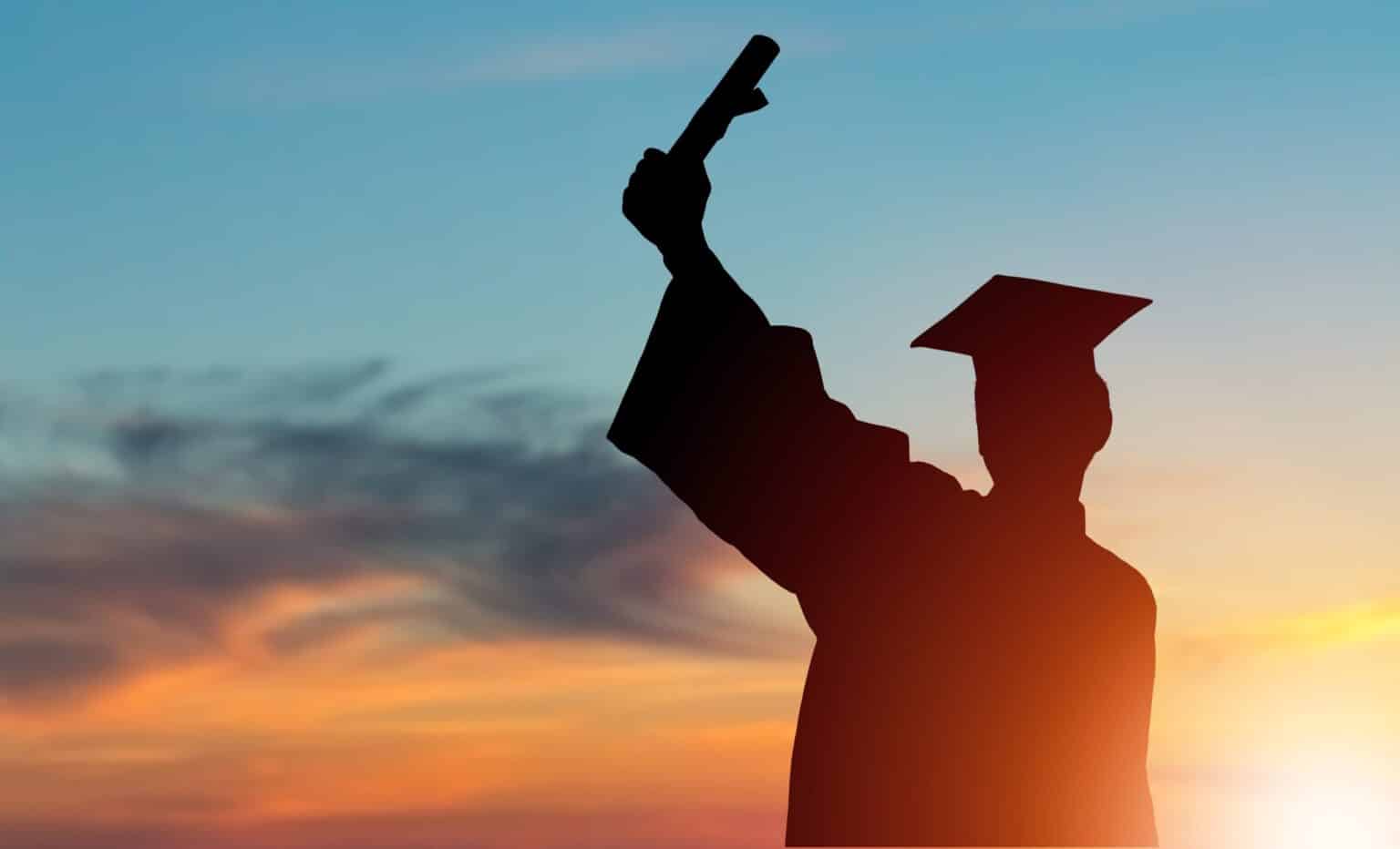 Silhouette of a graduate wearing a cap and gown holding up a diploma against a colorful sunset sky.