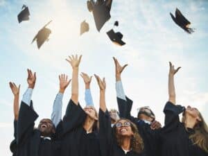 A group of graduates in caps and gowns celebrate by joyfully tossing their graduation caps into the air under a bright sky.