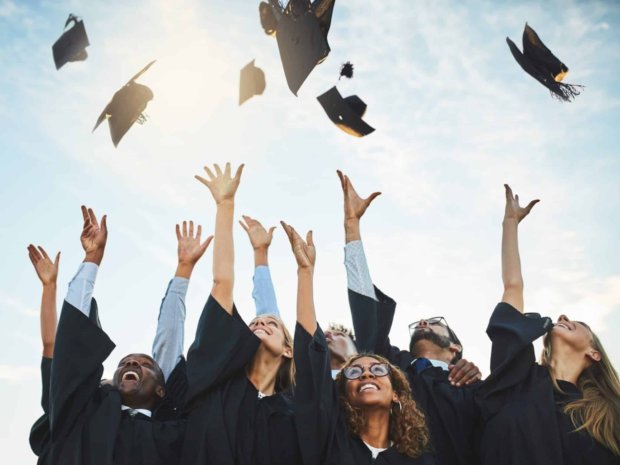 A group of graduates in caps and gowns celebrate by joyfully tossing their graduation caps into the air under a bright sky.