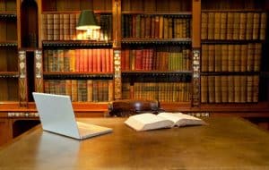 A wooden library table with an open book and a laptop sits in front of bookshelves filled with old, leather-bound books. A lamp above the shelves casts a warm light over the scene.