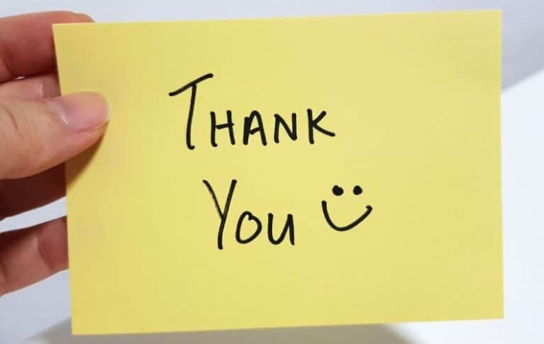 A hand holding a yellow sticky note with the words Thank You and a smiley face drawn in black marker.