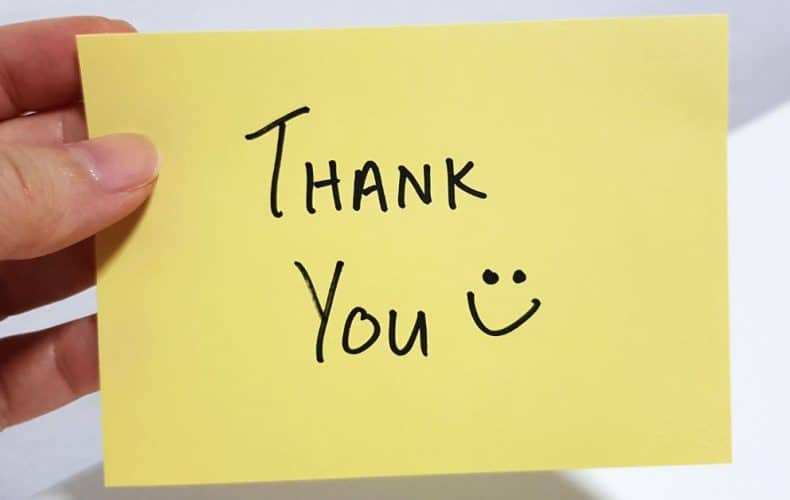 A hand holding a yellow sticky note with the words Thank You and a smiley face drawn in black marker.